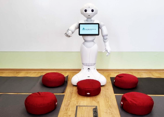 Roboter Pepper steht in einem Raum zwischen Matten und Sitzkissen. Er scheint auf Gäste zu warten und hält einladend den rechten Arm nach vorn.