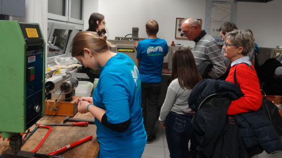 Das Bild zeigt den Blick in ein Labor der Werkstofftechnik an der EAH Jena. Die Mitarbeiter in blauen T-Shirts stehen an Geräten und an eienr Werkbank. Bei ihrer Arbeit schauen Gäste verschiedenen Alters interessiert zu.