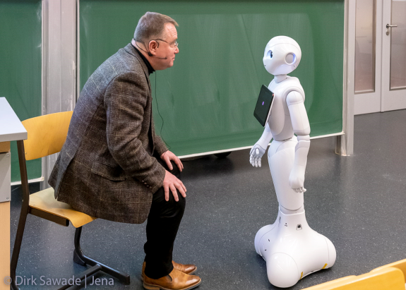 Das Bild ist während der Langen Nacht der Wissenschaften 2019 an der Ernst-Abbe-Hochschule Jena entstanden. Es zeigt Prof. Hundeck bei seinem damaligen Vortrag in der Interaktion mit seinem „Partner“ Pepper, einem humanoiden Roboter, der darauf programmiert ist, Menschen und deren Mimik und Gestik zu analysieren und auf diese Emotionszustände entsprechend zu reagieren. Die Künstliche Intelligenz spielt auch im diesjährigen Poetry Slam eine Rolle.