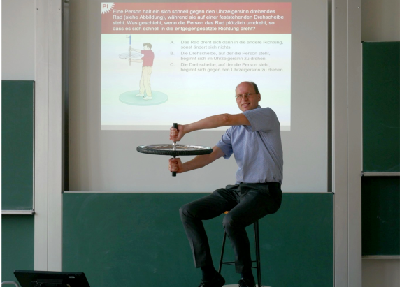 Prof. Sienz sitzt im Vordergrund, er hält ein Einrad mit beiden Armen und demonstriert den Versuchsaufbau einer seiner Quizfragen. Diese erkennt man im Hintergrund.