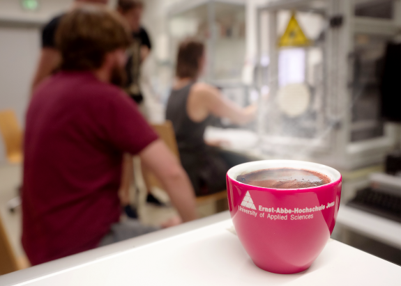 Im Vordergrund steht eine rote Kaffeetasse mit dem Aufdruck "Ernst-Abbe-Hochschule Jena". Im unscharfen Hintergrund erahnt man Personen der Arbeitsgruppe von Prof. Bliedtner.