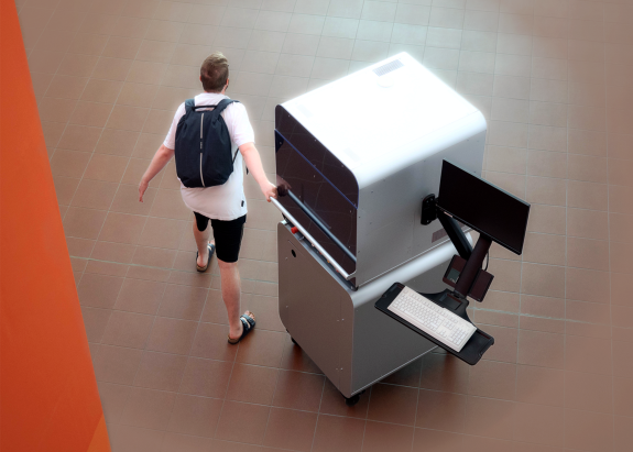 Ein Mann zieht den mobilen Laser (großes, geschlossenes Gerät, an der Seite außen erkennt man einen Monitor und eine Tastatur an einem Schwenkarm) hinter sich her durch ein Foyer der Hochschule.