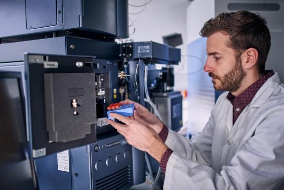 Ein Mann im Laborkittel stellt ein Gefäß mit mehreren Probengläschen in ein Analysegerät