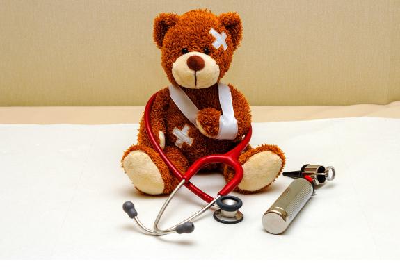 Auf einem Krankenbett sitzt ein Teddybär. Er hat zwei große Pflaster an Kopf und Bauch. Sein linker Arm ist in einer Schlinge. Um ihn geschlungen ist ein Stethoskop, rechts neben ihm liegt ein digitales Fieberthermometer.