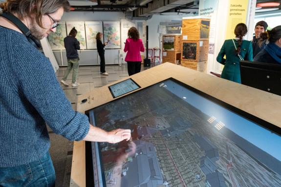 Ein Mann im Vordergrund interagiert mit einem großen, schrägen Touchscreen-Tisch, auf dem eine digitale 4D-Stadtkarte angezeigt wird. Im Hintergrund befinden sich weitere Personen in einem Ausstellungsraum mit verschiedenen Informationstafeln, Karten und Exponaten, die eine wissenschaftliche Ausstellung zeigen.