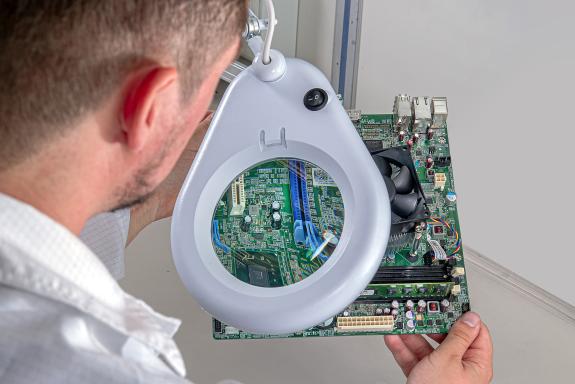 Ein Mann untersucht eine Computer-Motherboard durch eine große Lupe mit Licht und konzentriert sich auf die Schaltkreiskomponenten.