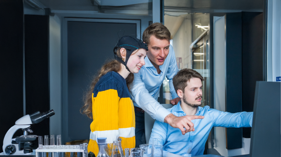 Drei Personen betrachten Forschungsergebnisse am Monitor; eine trägt eine Sensorkappe, daneben Mikroskop und Laborgefäße.