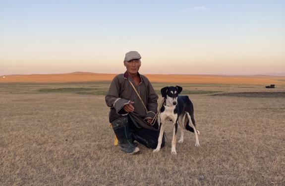 A man is walking a dog on a lead and is crouching beside it in a vast steppe landscape.