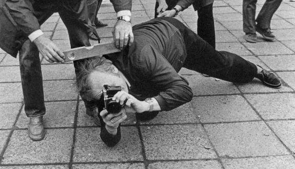 The black-and-white photograph shows a man lying in a public square with an analogue camera in front of his face. He is accompanied by other people. One person is holding a spirit level. 