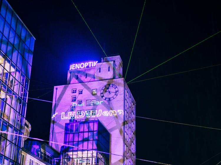 Blick auf das Jenaoptik Hochhaus, es wird mit liala Licht angestrahlt. Zudem wird mit Laserlicht der Text LNDW Jena drauf gestrahlt.